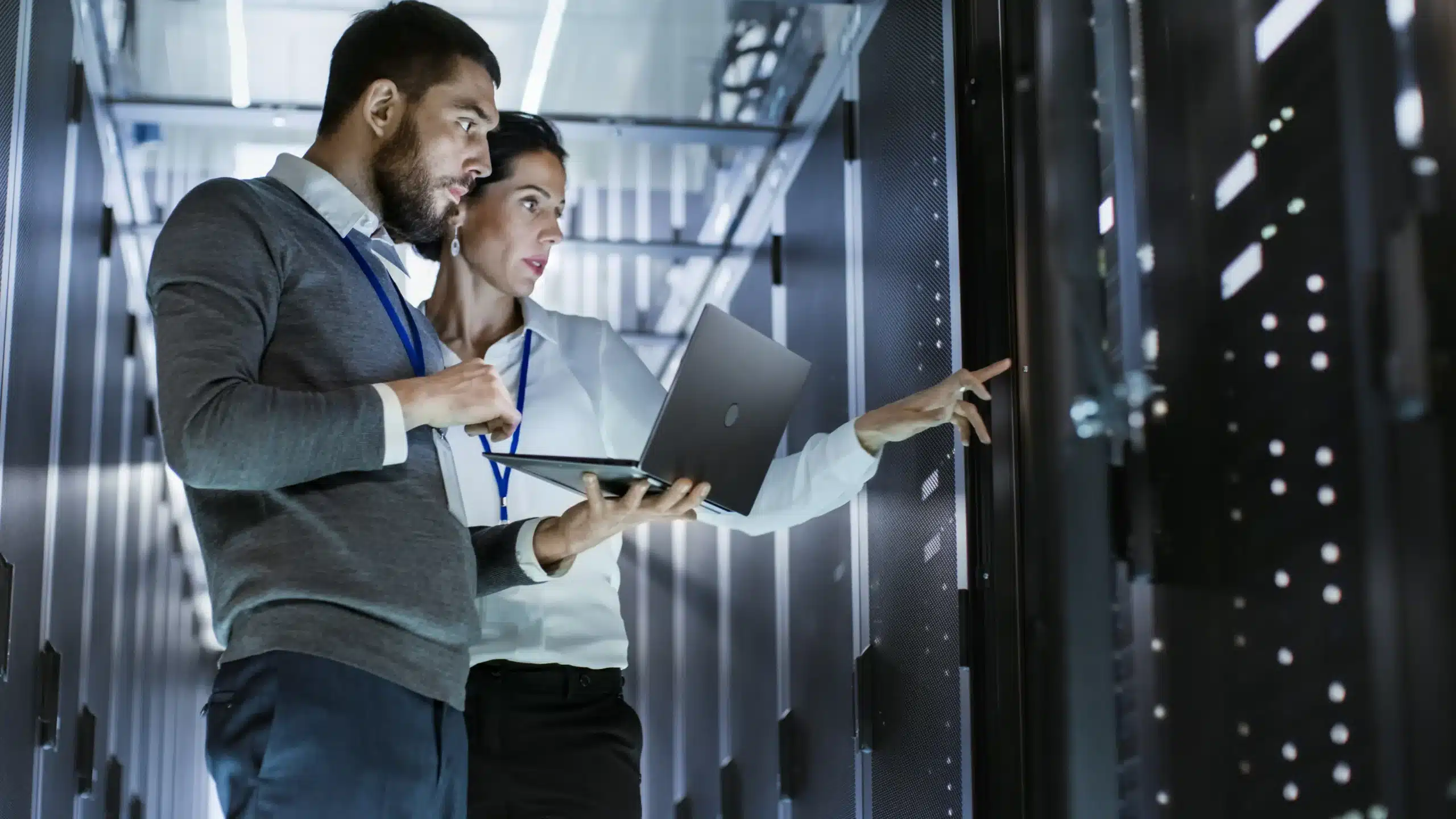 Two professionals in a server room discussing data while examining a server rack, one holding a laptop and the other pointing at the equipment.