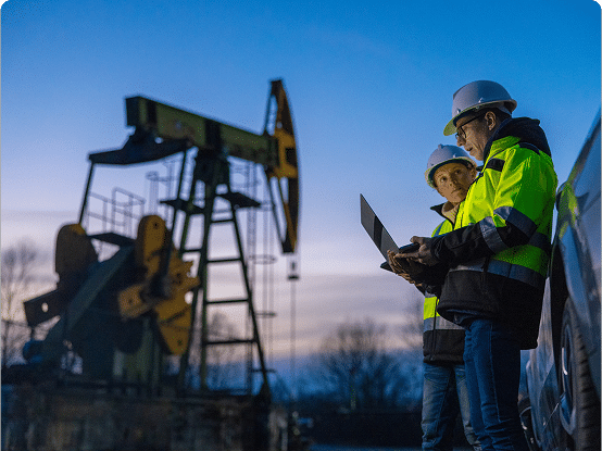 Two workers in safety gear near an oil pump jack at twilight, one holding a laptop.
