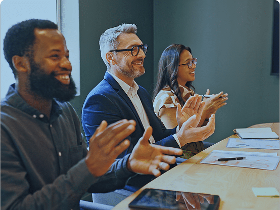 Three people sitting at a table clapping, with papers, a pen, and a tablet in front of them.