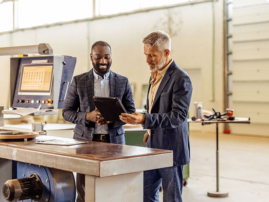 Two professionals in an industrial setting reviewing data on a tablet near machinery.