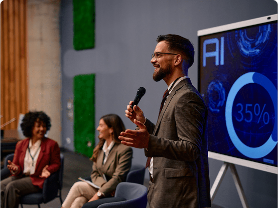 Three people in a professional setting with one speaking into a microphone and a screen displaying "AI" and "35%" with related graphics.