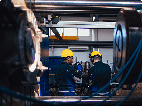 Two workers in yellow hard hats and blue uniforms in an industrial setting with pipes and metal structures