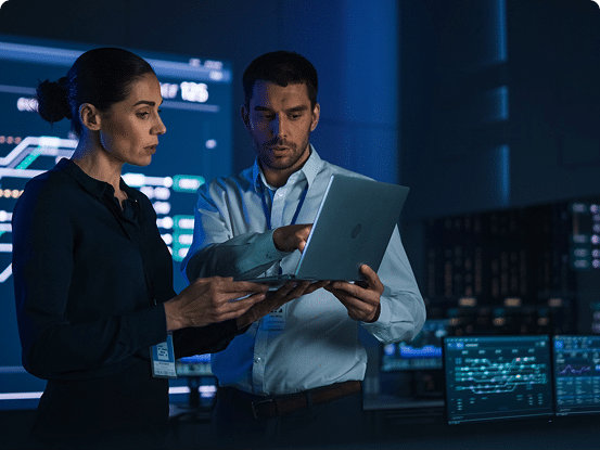 Two people in a dimly lit room viewing a laptop, with multiple computer screens showing data and graphs in the background.