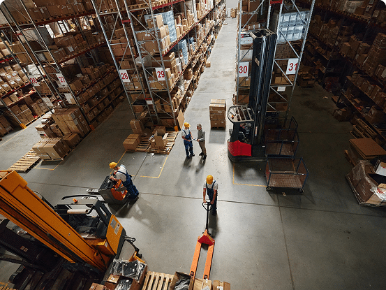 Warehouse with tall shelves, labeled sections, and workers in safety gear operating forklifts and pallet jacks