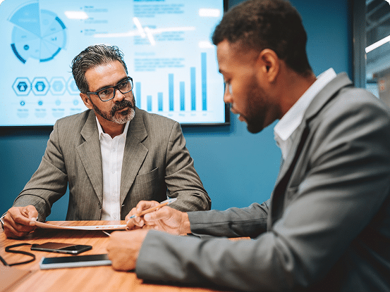 Two individuals in suits at a business meeting table with documents and devices, with charts and graphs displayed on a large screen in the background.