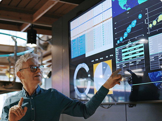 Person pointing at a large screen with charts, graphs, and tables in an indoor, industrial setting.