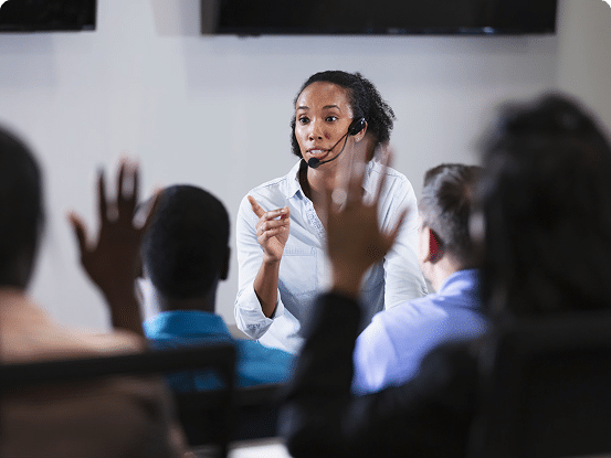 Person speaking to a group in a classroom or seminar setting, with some audience members raising their hands.