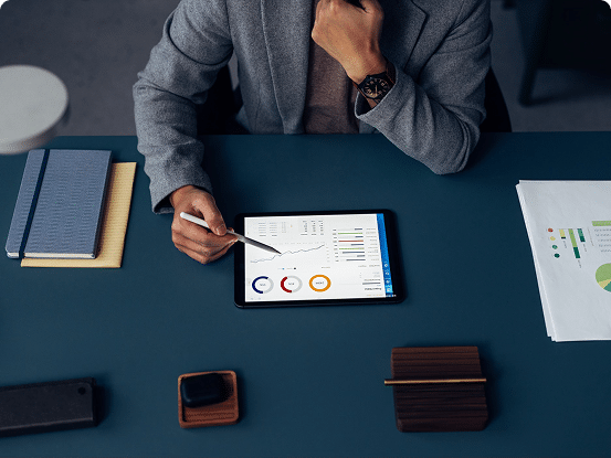A person uses a stylus on a tablet displaying charts and graphs, with notebooks, a smartphone, and documents spread across the desk.