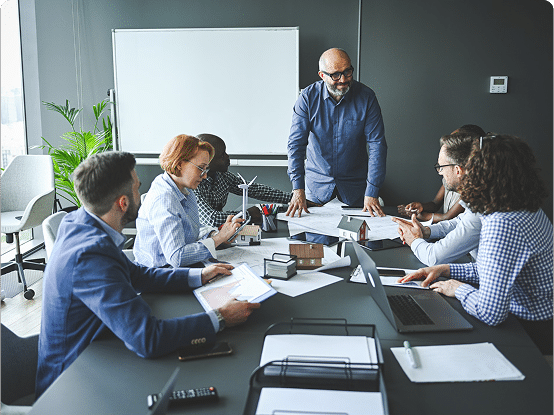Six people in a meeting room gathered around a table with documents and laptops; one person stands leading the discussion, with a whiteboard and plants in the background.
