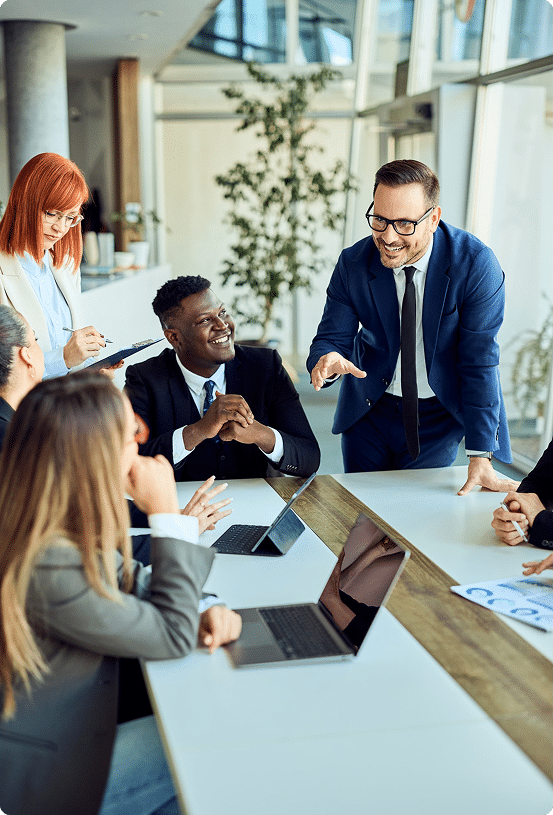 Business meeting with six people in an office, one man standing and presenting to others seated around a table with laptops and documents