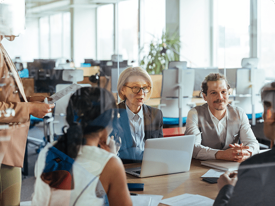 A group of people in a modern office sit around a table with papers and a laptop, while one person stands presenting with documents in hand.
