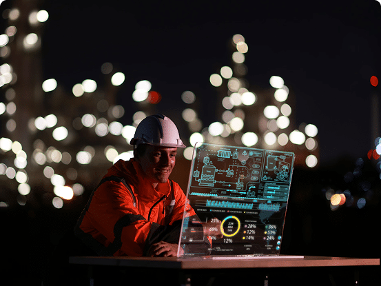 Person in orange jacket and white hard hat viewing data on a transparent screen in a brightly lit industrial setting at night.