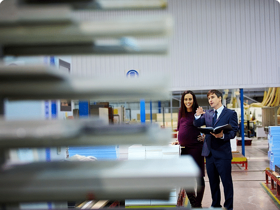 Two people stand in a factory or warehouse; one gestures while holding an open book, surrounded by shelves of materials and industrial equipment.