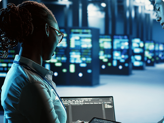Person in a server room using a laptop and tablet, surrounded by server racks.