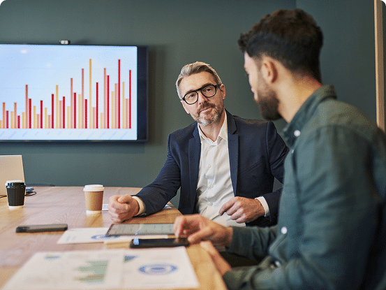 Two people in a meeting room with documents, devices, and a bar graph on a screen in the background.