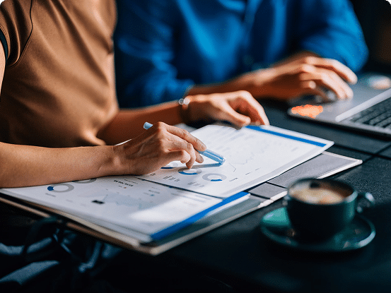 Two people working at a table; one points at a document with charts while the other types on a laptop.