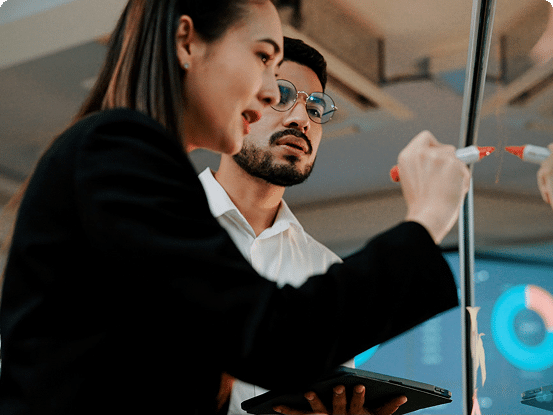 Two professionals in business attire collaborate at a glass board; one writes with a red marker while the other holds a tablet, with charts and graphs visible in the background.