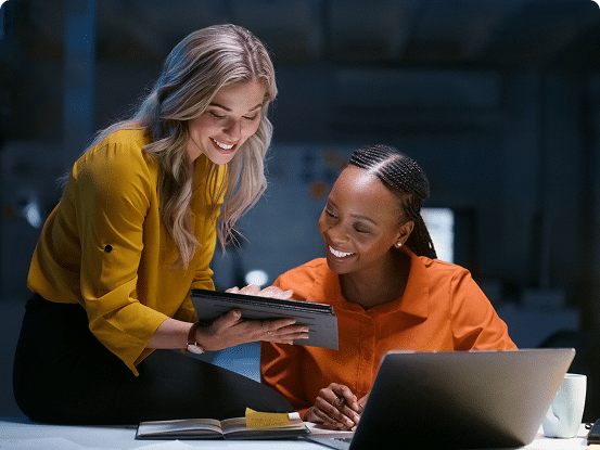 Two people in a professional setting, one standing with a tablet and the other sitting at a desk with a notebook and laptop.