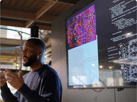 A person presents in front of a large screen displaying technical diagrams, a circuit board layout, and lines of code.