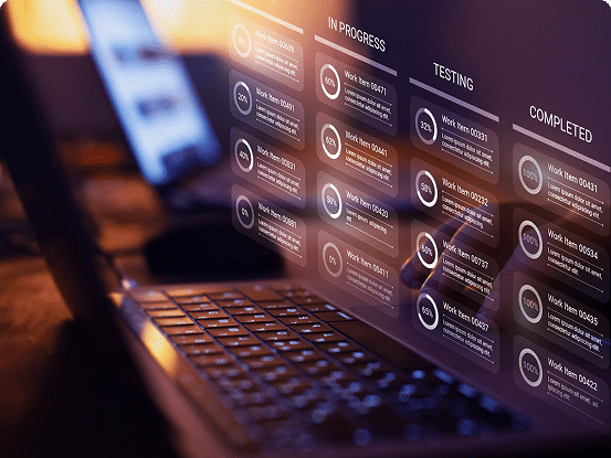 Laptop screen showing a project management interface with columns for "In Progress," "Testing," and "Completed" work items and progress percentages.