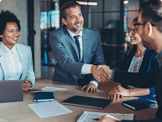 Four people in an office setting, with two shaking hands across a table covered in documents and a laptop.