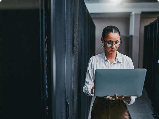 Person working on a laptop in a server room with tall black server racks on both sides.