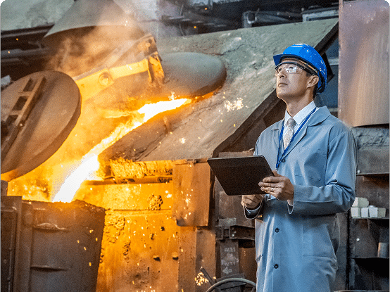 Person in safety gear holding a clipboard near machinery pouring molten metal in an industrial setting.