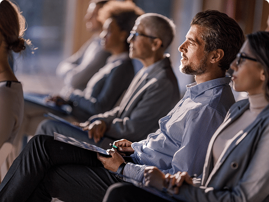 Group of people sitting in a row at a seminar or conference, with blurred faces and some holding notebooks or documents.