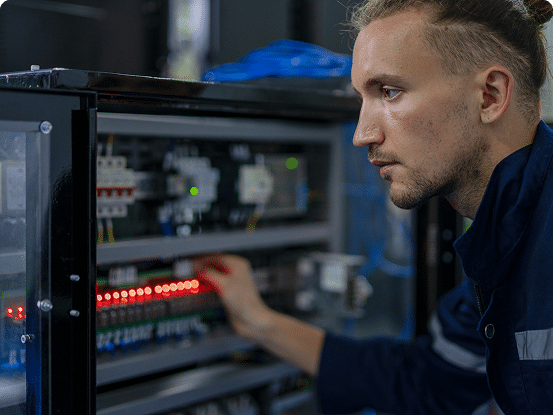 Person inspecting an electrical control panel with circuit breakers, relays, wiring, and illuminated red indicator lights.