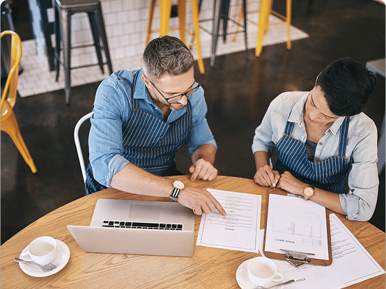 Two people in aprons discussing documents at a café table with a laptop and coffee cups.