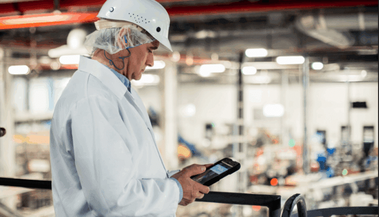 Person in lab coat and hard hat using a tablet in an industrial facility.