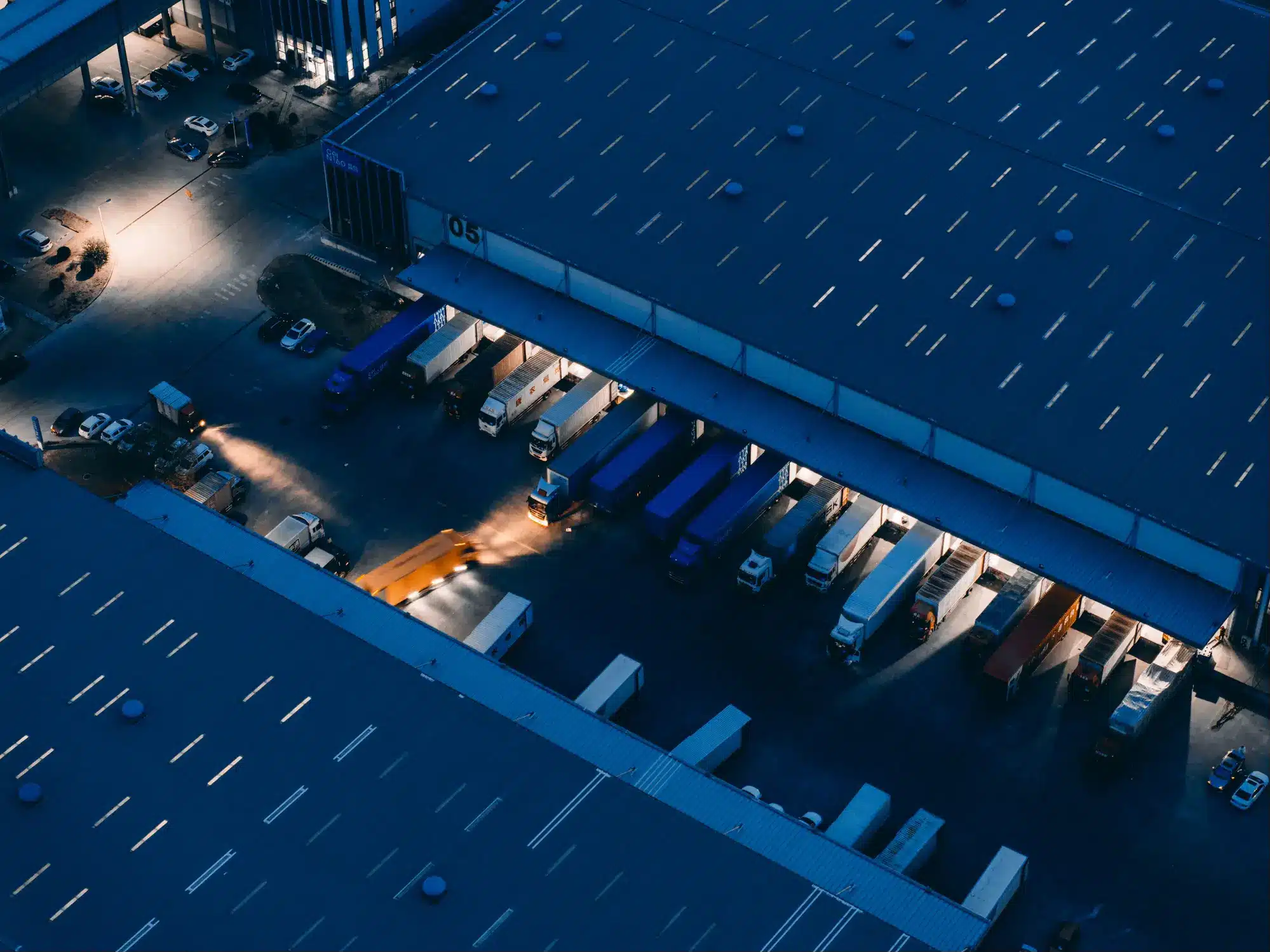Aerial night view of an industrial warehouse with trucks at loading docks under artificial lighting.