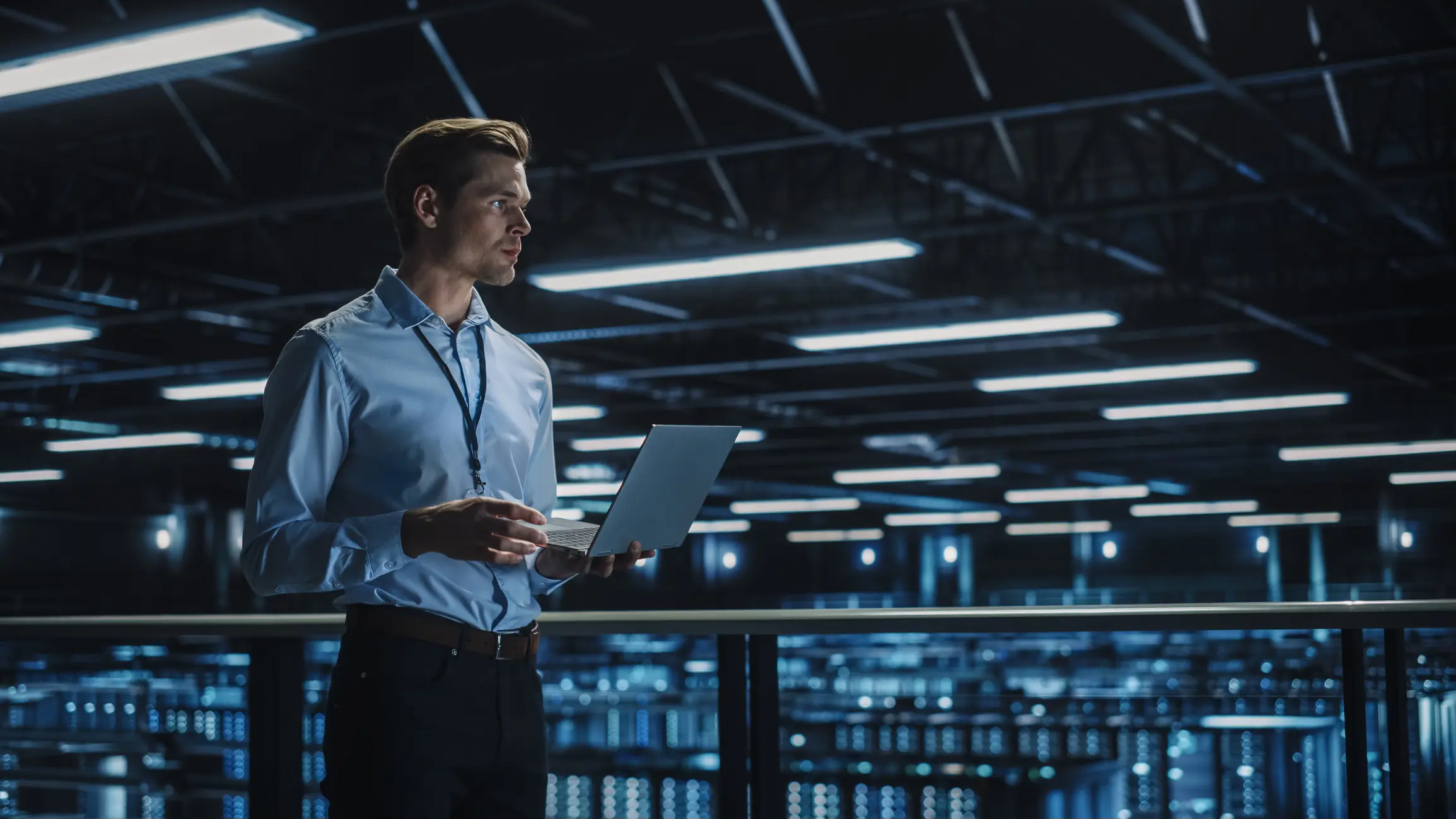 A professional in a blue shirt stands in a high-tech data center, holding a laptop and looking thoughtfully at the screens.