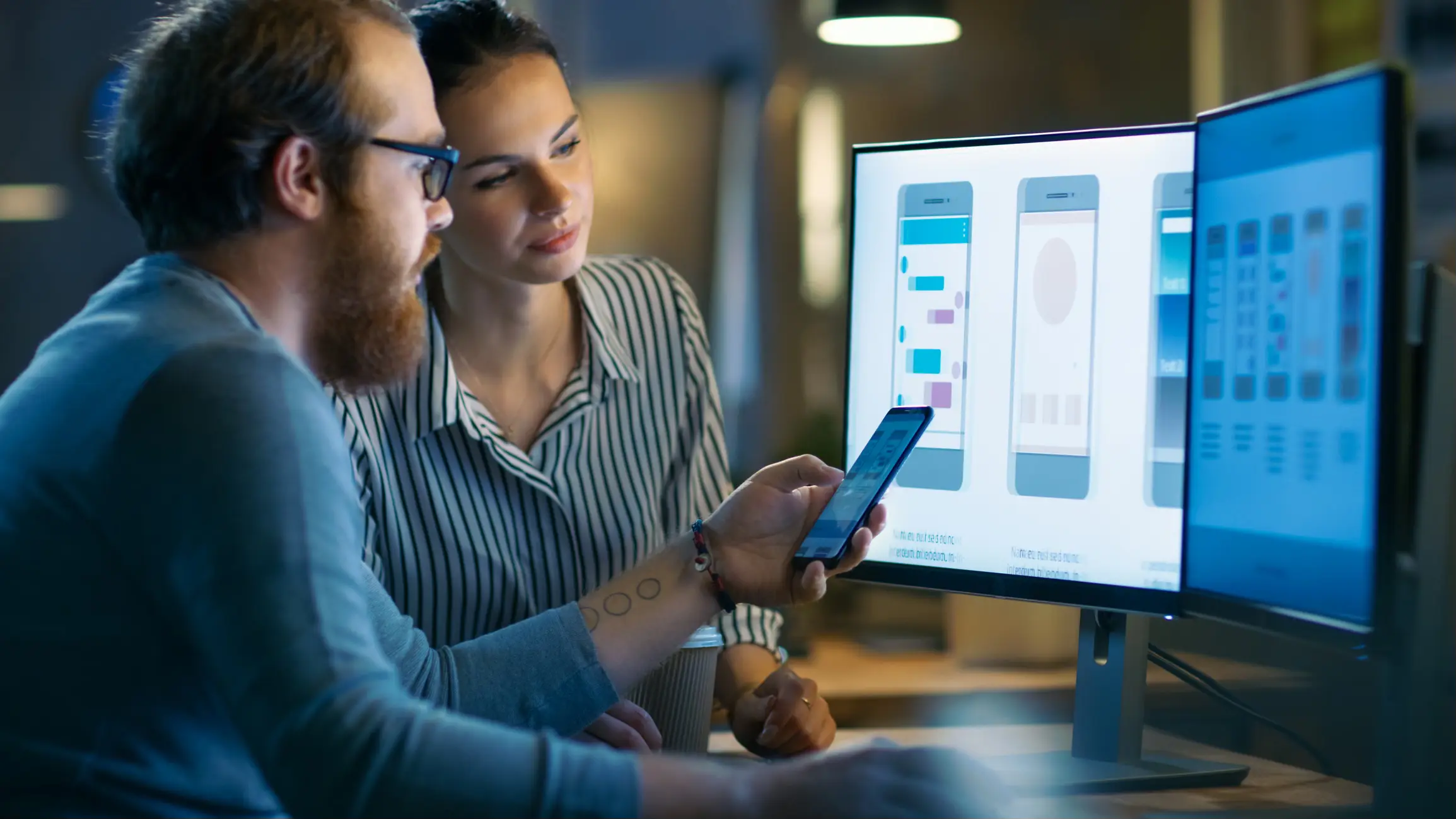 A person holds a smartphone while discussing app designs displayed on two computer monitors in a modern workspace setting.