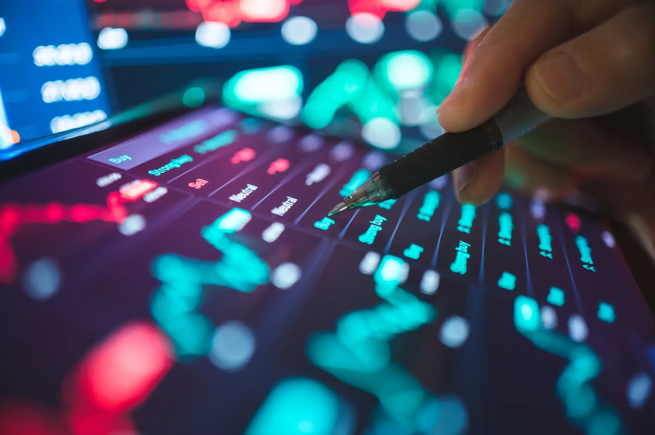 A hand holds a pen above a tablet displaying vibrant stock market data with 'Buy' and 'Sell' options highlighted.